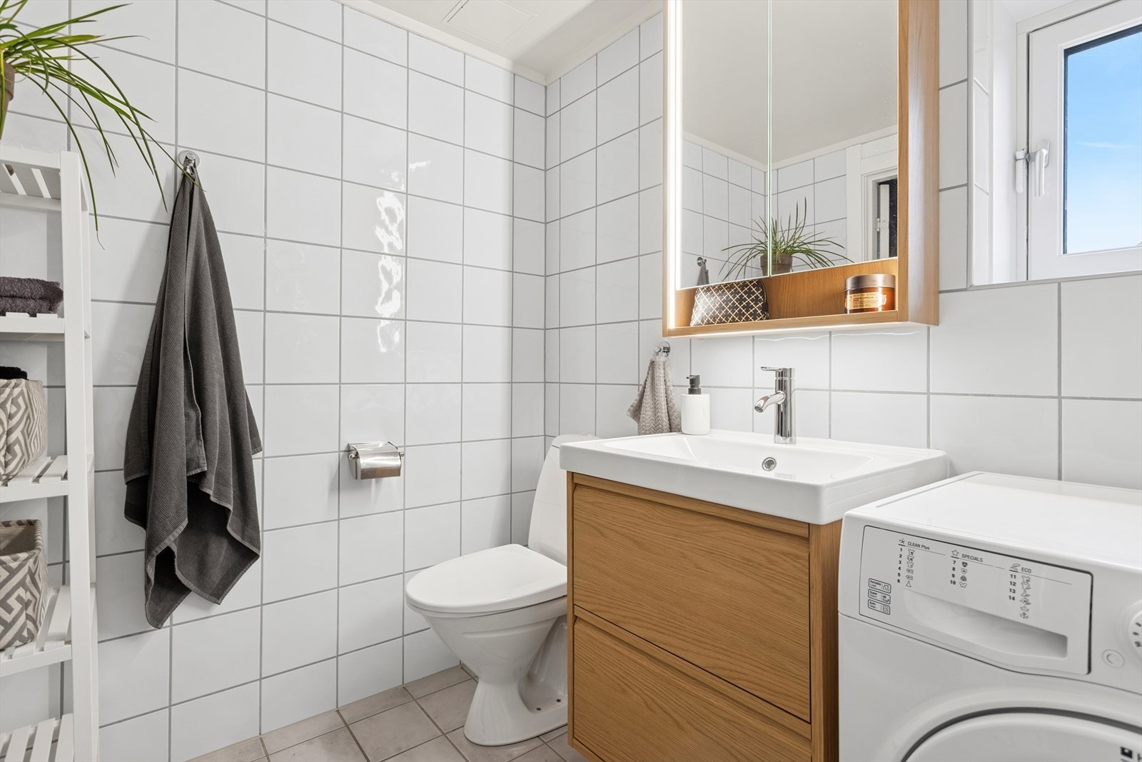 Badet er utstyrt med gulvstående toalett og dusjhjørne, samt en stilren servantinnredning med heldekkende servant, skuffefronter i finer og tilhørende speilskap med integrert belysning. Baderomsinnredningen var ny i 2025. Galleribilde