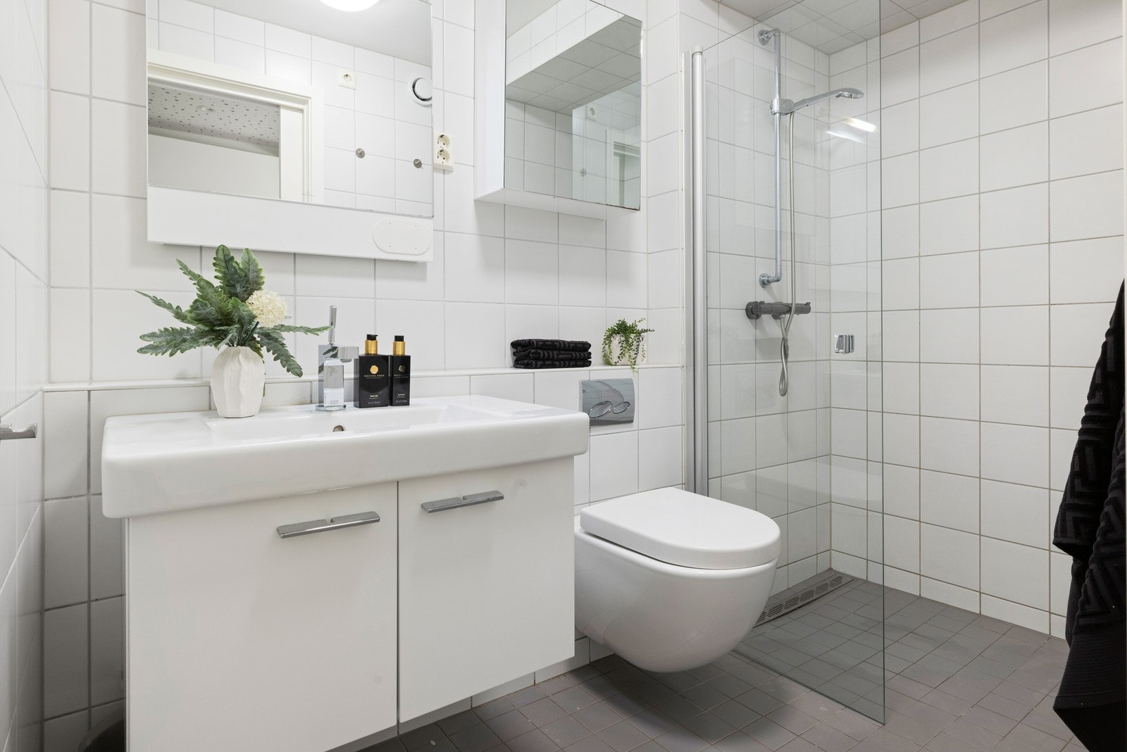 Flislagt bad innredet med servantskap med servant og ettgreps blandebatteri og vegghengt klosett samt dusjhjørne med nisjedører i herdet glass Galleribilde