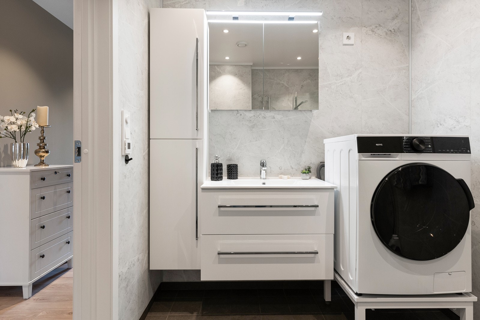 Bad i 1. etasje har grå fliser med varmekabler i gulv, baderomsplater med marmormøster og slett hvit tak med spotter. Hvit baderomsinnredning med skuffer og heldekkende servant. Speilskap og belysning. Høyskap for oppbevaring. Opplegg for vaskemaskin. Galleribilde