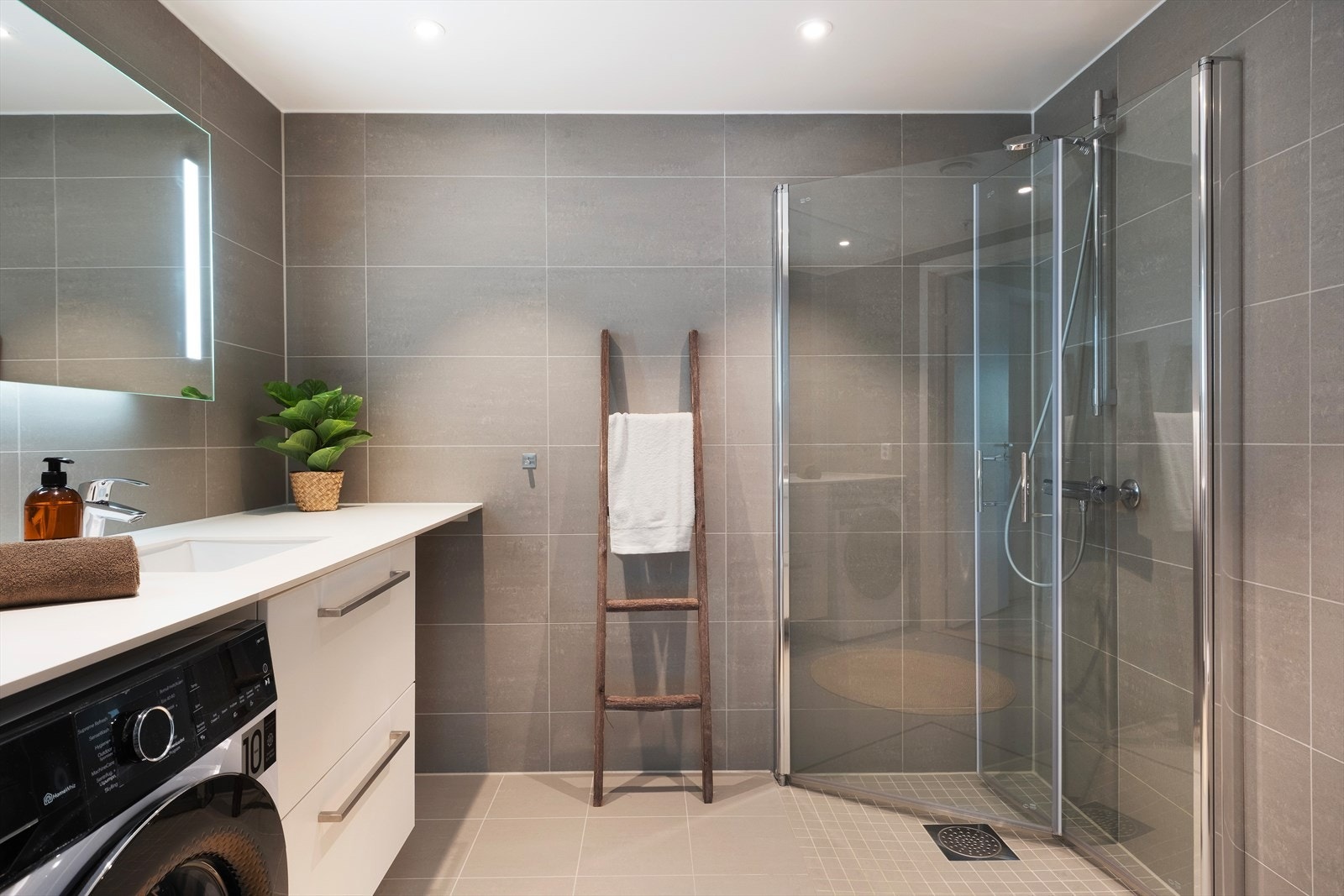 Badet har delikat og stilfull innreiing med nedfelt servant med underskuffer, benkeflate med opplegg for vaskemaskin og tørketrommel, dusjhørne med innfellbare glasveggar og vegghengt toalett. Galleribilde