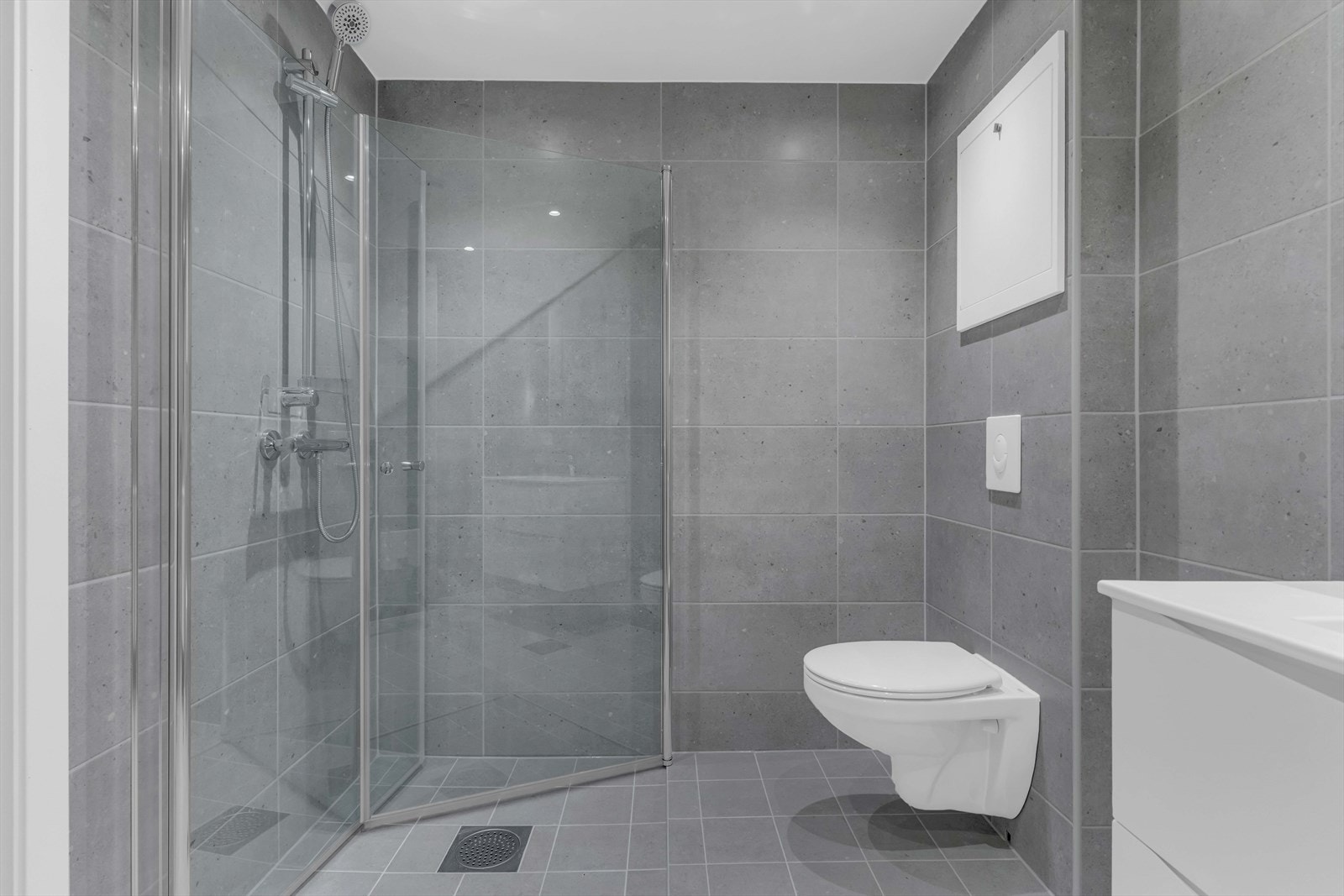 På badet er det en hvit, moderne skuffeseksjon med heldekkende servant, og over er det et dobbelt speilskap med belysning. Videre har badet et vegghengt toalett og et dusjhjørne med innfellbare glassdører. Galleribilde