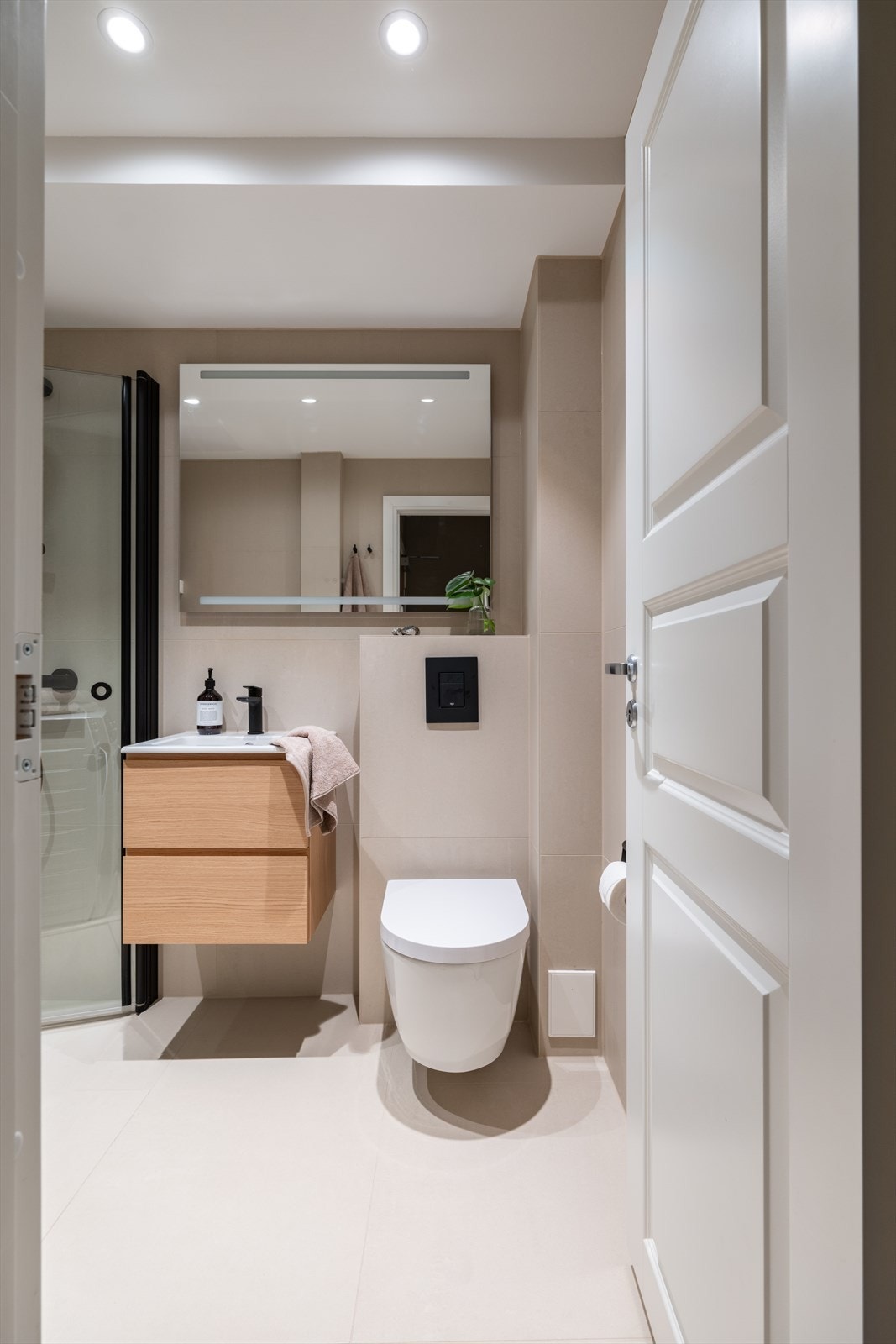 Videre er badet utstyrt med servant over skuffe-innredning, speil med sminkelys, veggmontert toalett og dusjhjørne. Galleribilde