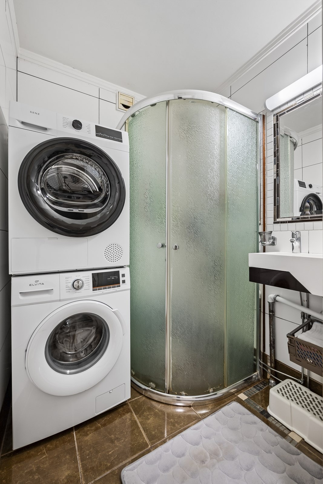 Flislagt baderom som inneholder dusjhjørne, servant, vaskemaskin og tørketrommel. Galleribilde