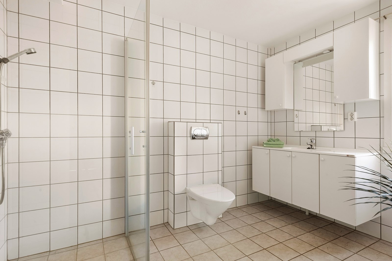 Romslig flislagt bad. Vegghengt toalett og dusjhjørne. Innredning med heldekkende servant og skap. Galleribilde