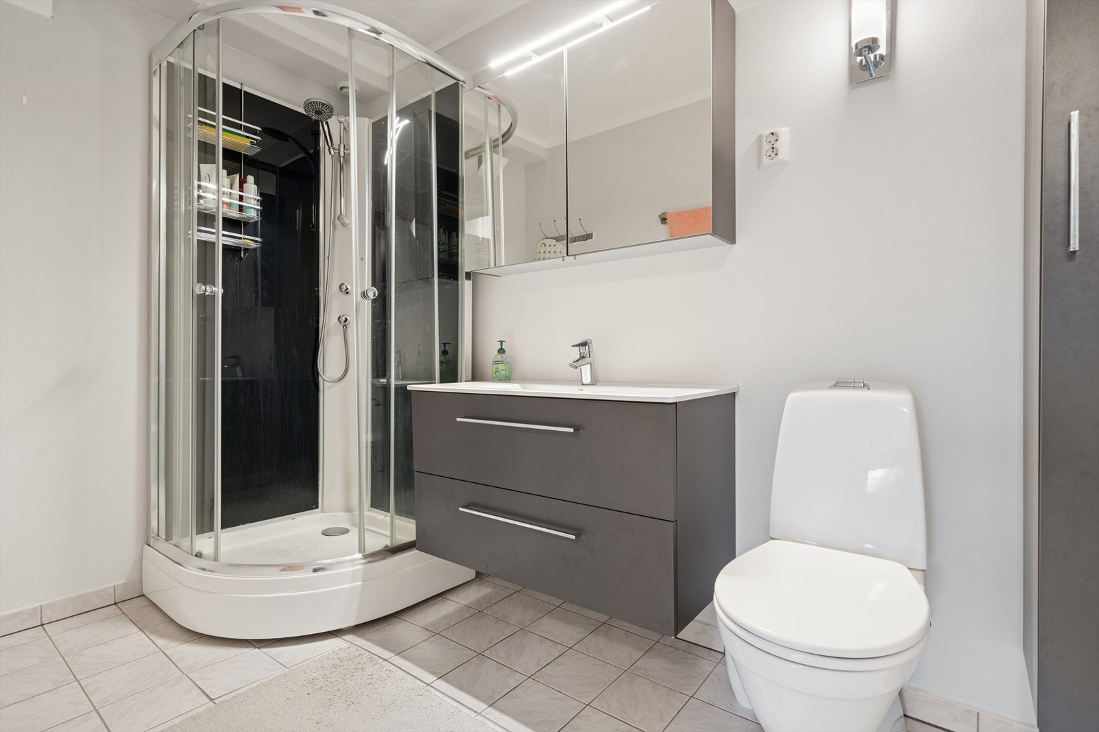 Lyst bad med flislagt gulv og varmekabler. Badet er utstyrt med dusjkabinett, toalett og en stilren baderomsinnredning. Galleribilde