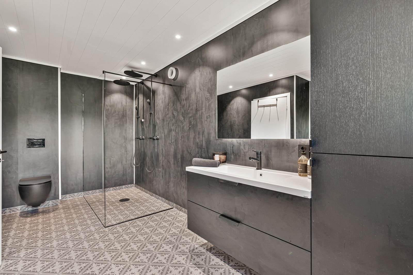 Stilfullt bad/vaskerom med gulvbelegg med varmekabler, baderomsplater på vegg og panel i tak.
Rommet er innredet med dusjhjørne med glassdør, dobbeldusj med regndusjer, vegghengt toalett og baderomsinnredning med vask, skuffeseksjon, skap og speil. Galleribilde