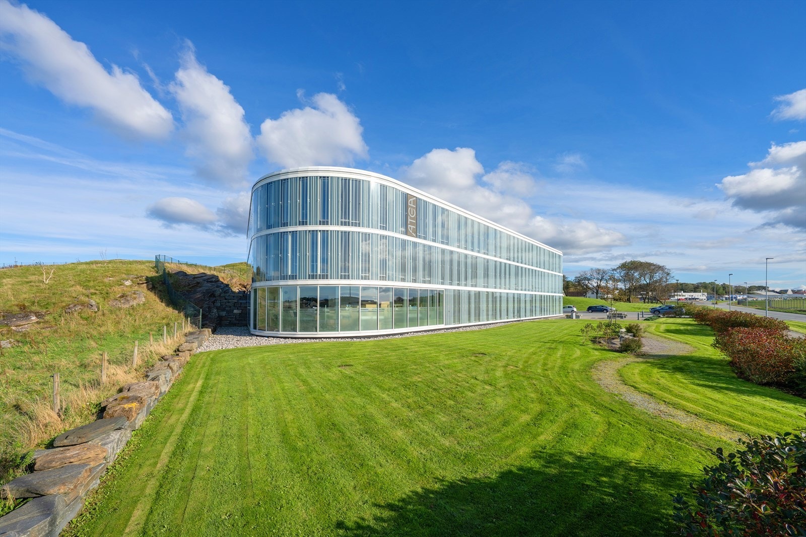 Moderne, buet bygning med glassfasade, omgitt av frodig grøntareal. Naturskjønn beliggenhet med enkel tilgang til vei og fasiliteter. Galleribilde