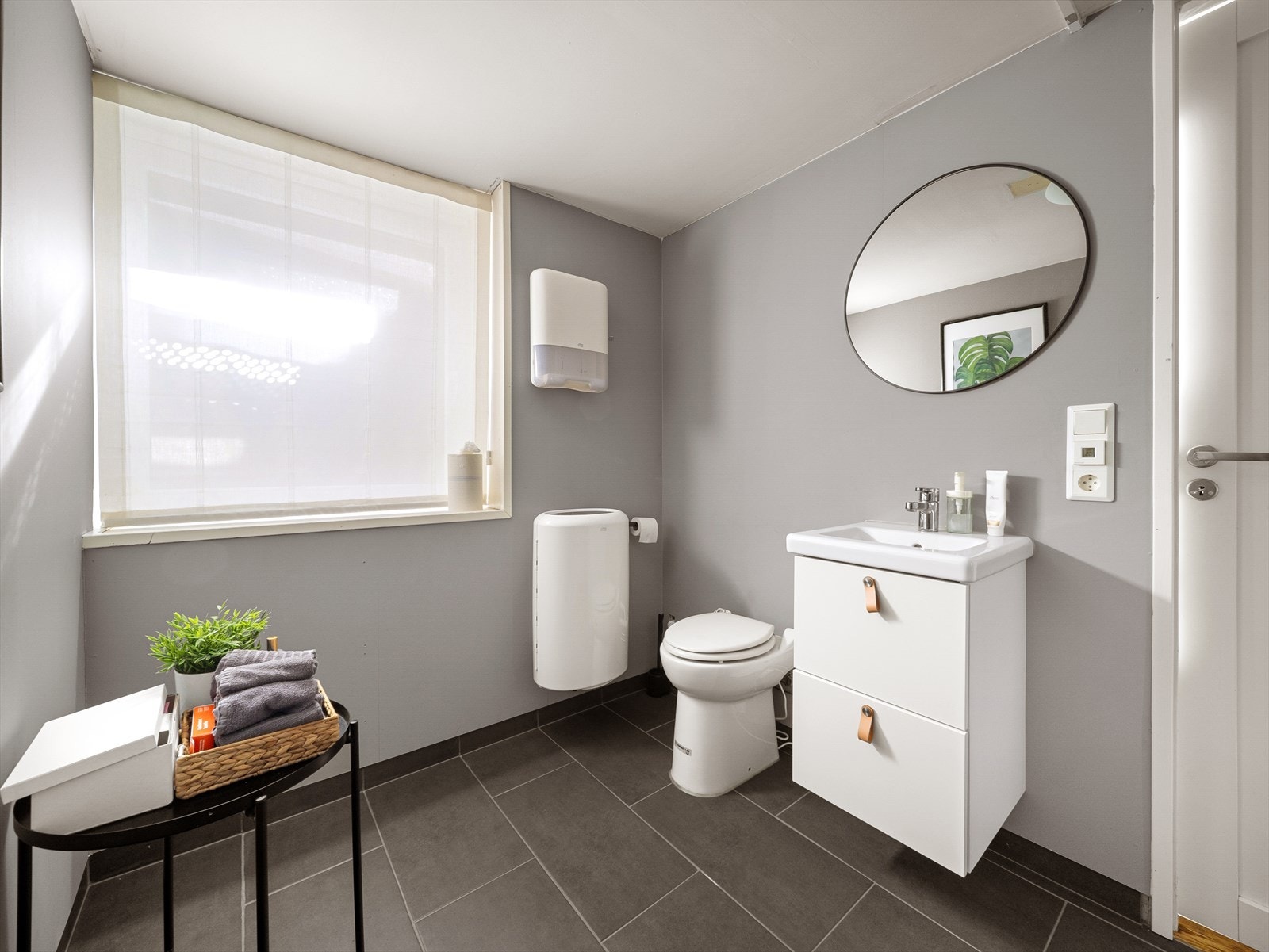 Toalettrom med fliser på gulv, malt gips på vegg, toalett, avløp fra toalett med kvern og pumpe, vask med underskap og lys i tak. Galleribilde