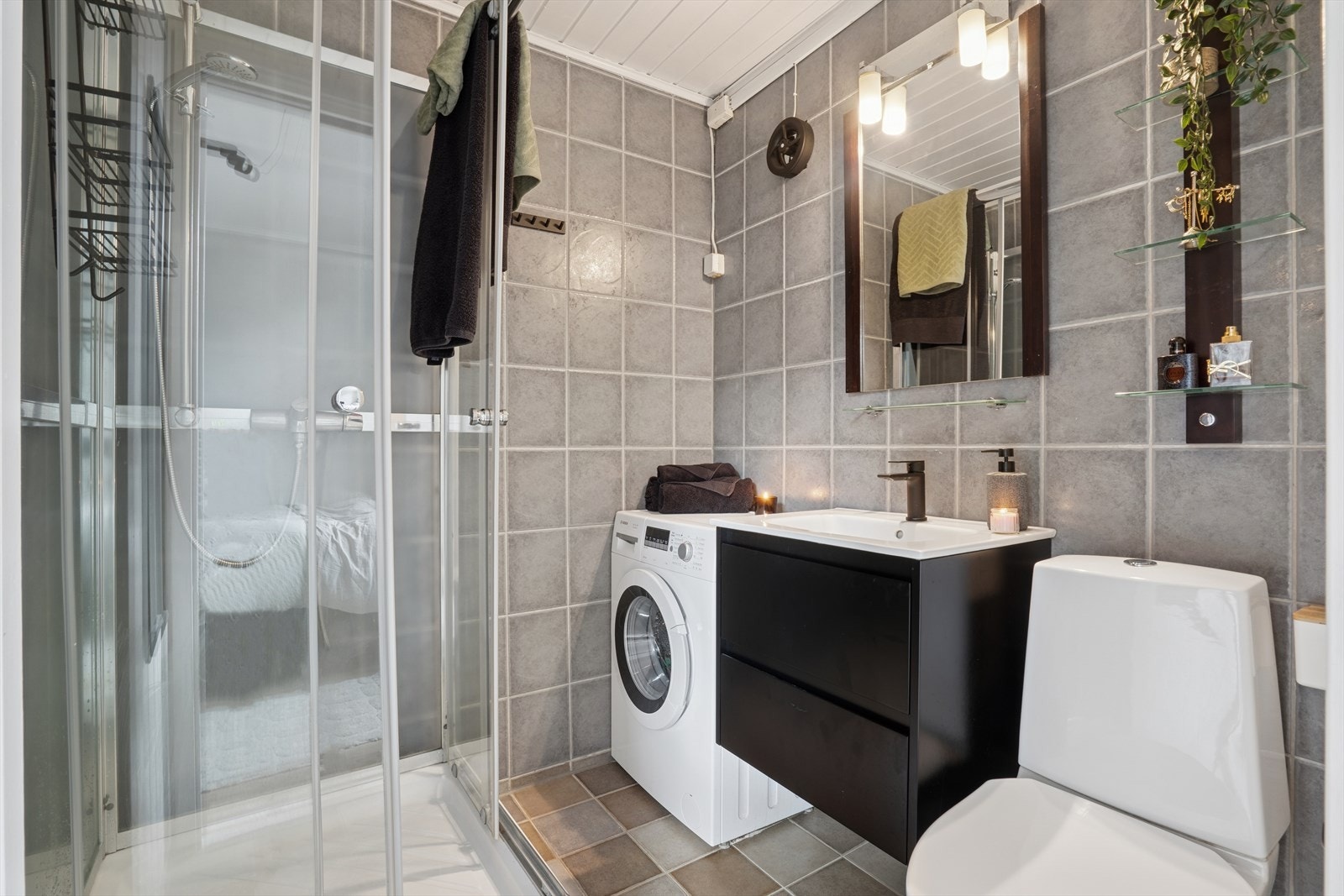 Pent flislagt baderom på ca. 3 m² med malt panel i himling. Badet er utstyrt med dusjkabinett, gulvmontert toalett, innredning med servant og opplegg for vaskemaskin. Galleribilde