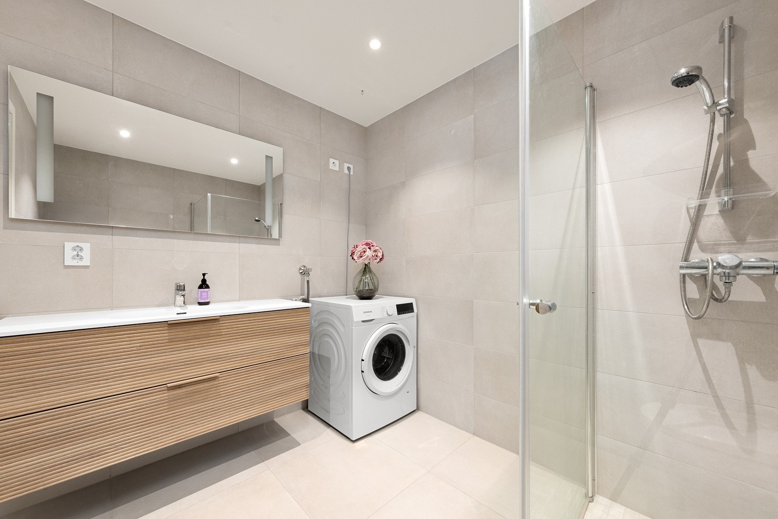 Det er store skuffer til oppbevaring under servant, speil med lys, lyse fliser på gulv og vegger, toalett og dusjvegger/hjørne. Galleribilde