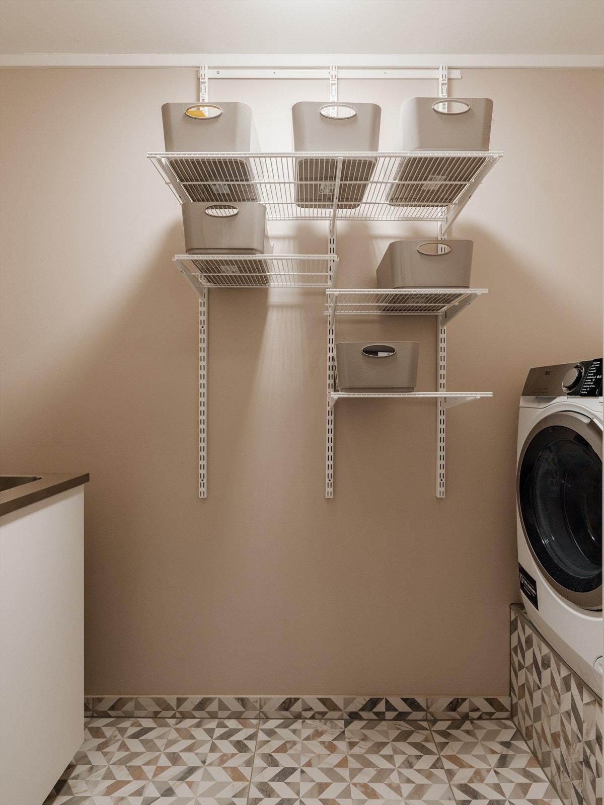 Praktisk vaskerom etablert i 2021, med flislagt gulv, åpne hyller og opplegg for vaskemaskin. Galleribilde