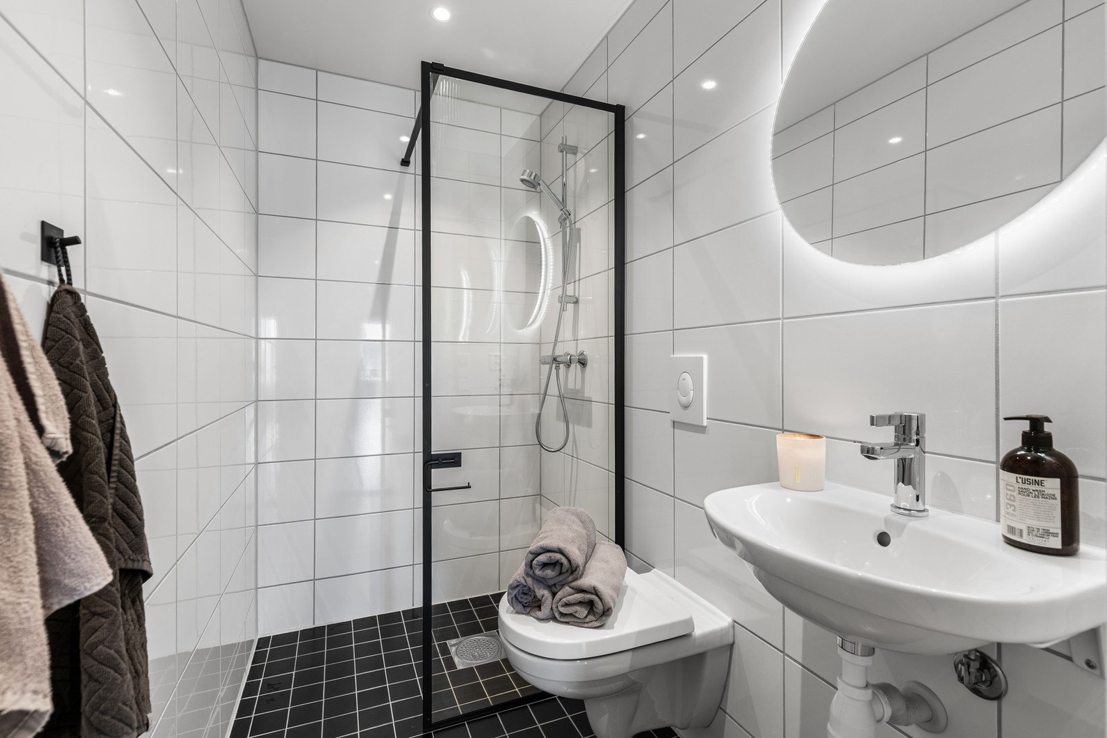 Baderom nr. 2. Dette badet har et dusjhjørne med en glass vegg, enkel servant og et tilhørende rundt speil med lys bak. Galleribilde