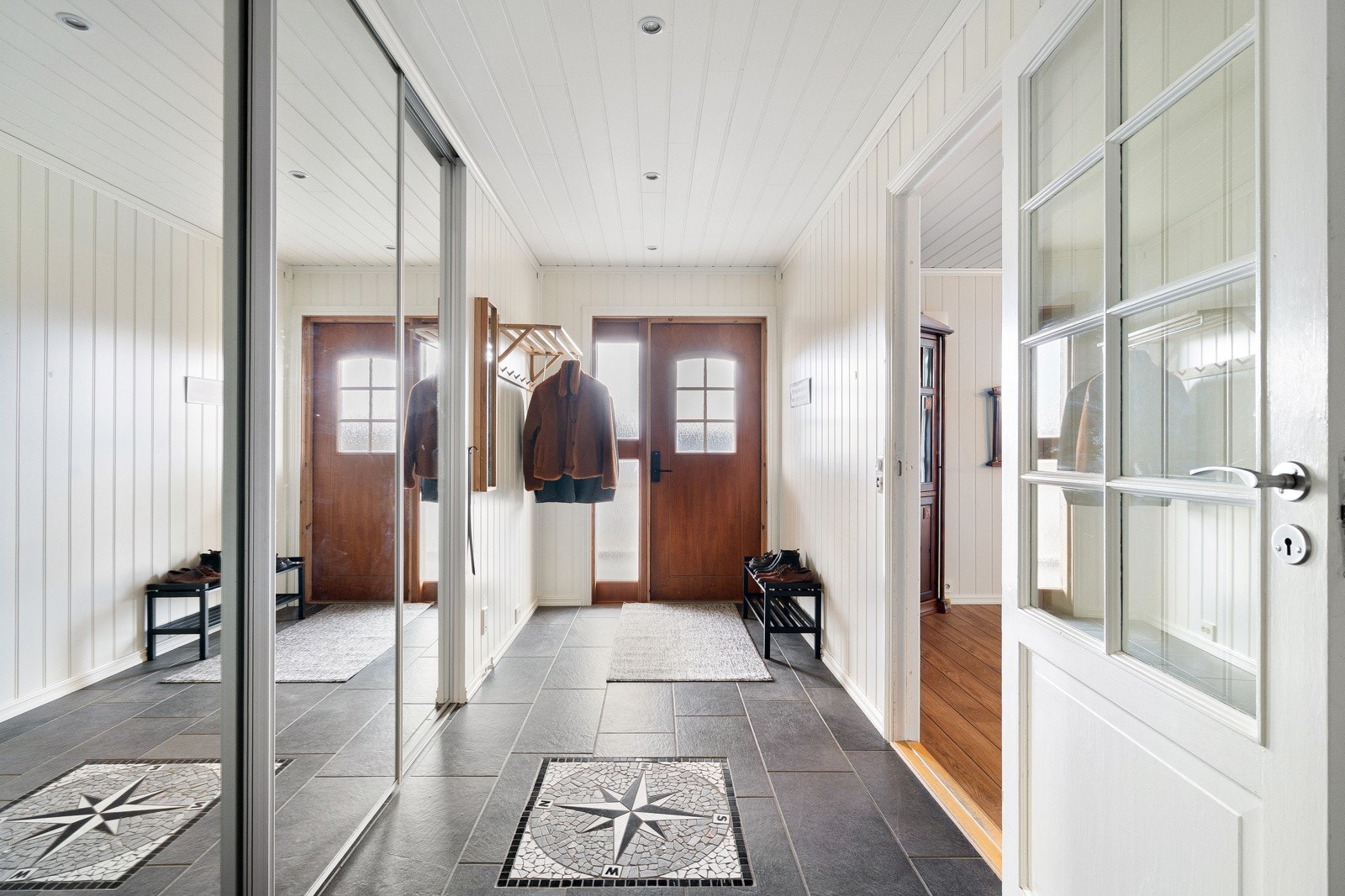 Flislagt inngangsparti med god plass for å henge fra seg yttertøyet i lekker skyvedørsgarderobe med speil.