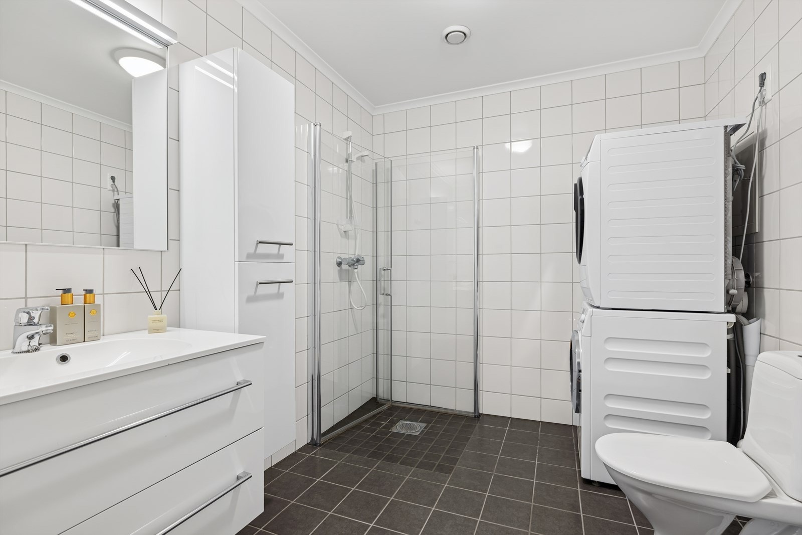 Bad utstyrt med toalett, dusjhjørne, opplegg til vaskemaskin og tøketrommel, servant og skap Galleribilde