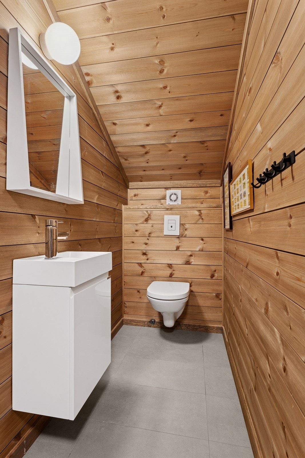 Separat toalettrom med vegghengt toalett og en vask med underskap. Overflatene består av panel på vegger og i tak, samt fliser på gulvet. Galleribilde