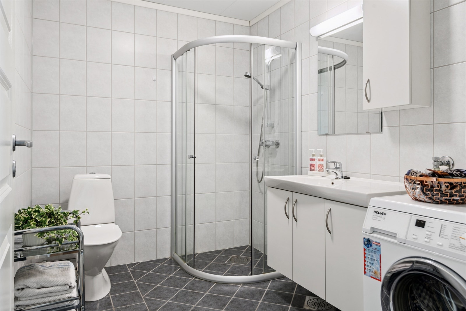 Flislagt bad med dusjhjørne, servantinnredning og opplegg for vaskemaskin. Galleribilde
