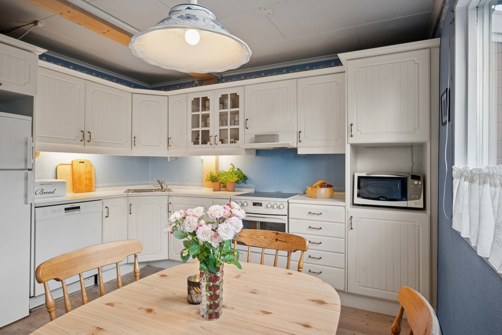 Kjøkkeninnredning med profilerte fronter og enkelte glassfronter i overskap. Det er belysning under overskap og en laminat benkeplate med nedfelt kum i rustfritt stål. Galleribilde