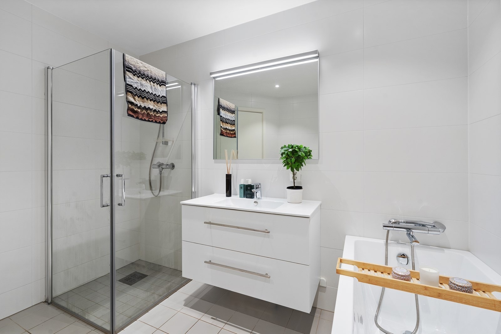 Badet er utstyrt med heldekkende servant over skuffeinnredning, overliggende spil med belysning, vegghengt toalett, badekar og dusjhjørne med innfellbare glassvegger Galleribilde