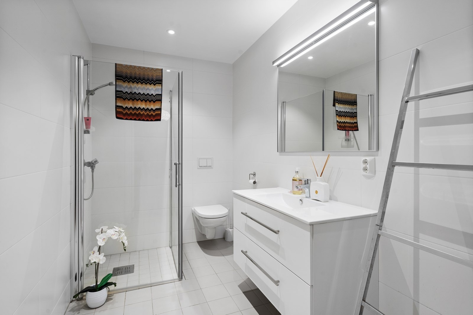 Lyst og tiltalende flislagt baderom med varmekabler i gulv. Baderommet er utstyrt med heldekkende servant over skuffeinnredning, overliggende spil med belysning, vegghengt toalett og dusjhjørne med innfellbare glassvegger Galleribilde