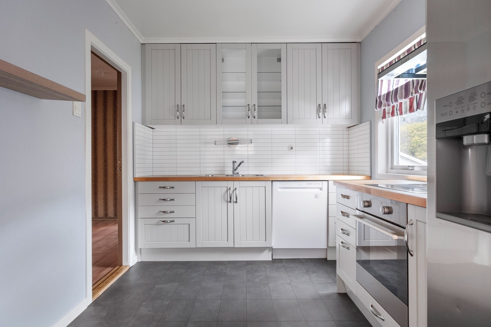 Kjøkkenet har innredning med profilerte fronter fra 2000-tallet. Benkeplaten er av heltre, flisplate på vegg over kjøkkenbenk. Det er side by side kjøl/fryseskap, oppvaskmaskin og integrert platetopp og stekeovn. Galleribilde