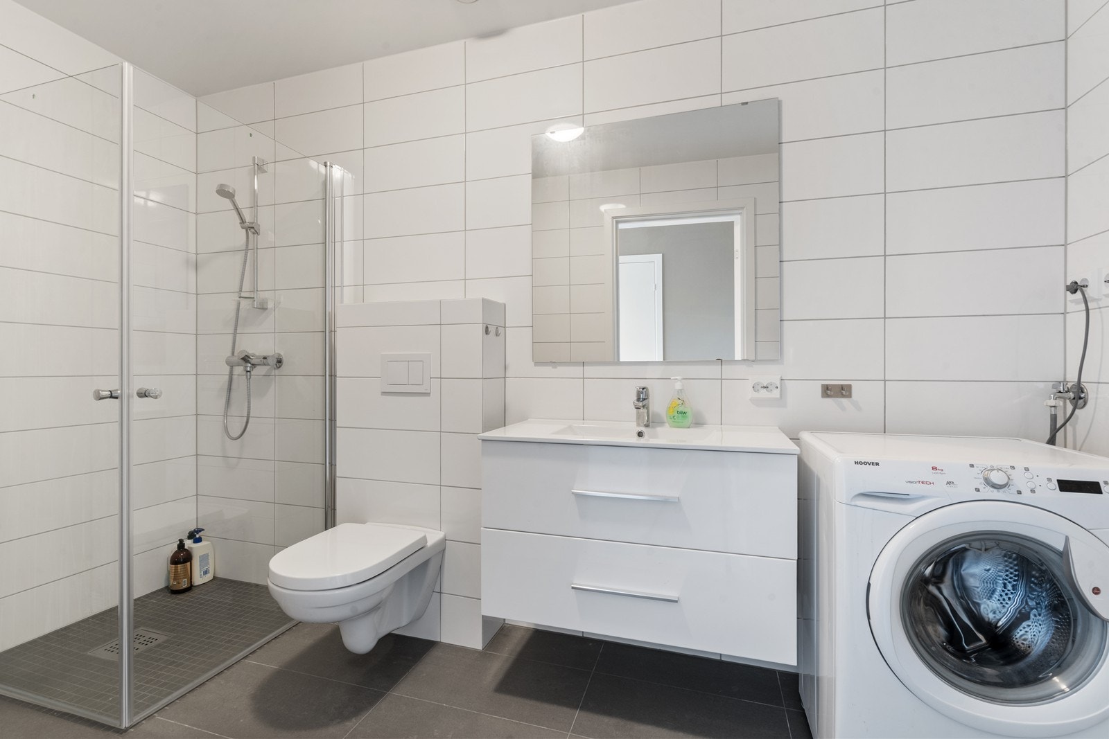 Lyst, helfliset bad med vegghengt toalett, hvit baderomsinnredning og dusjnisje med glassvegger Galleribilde