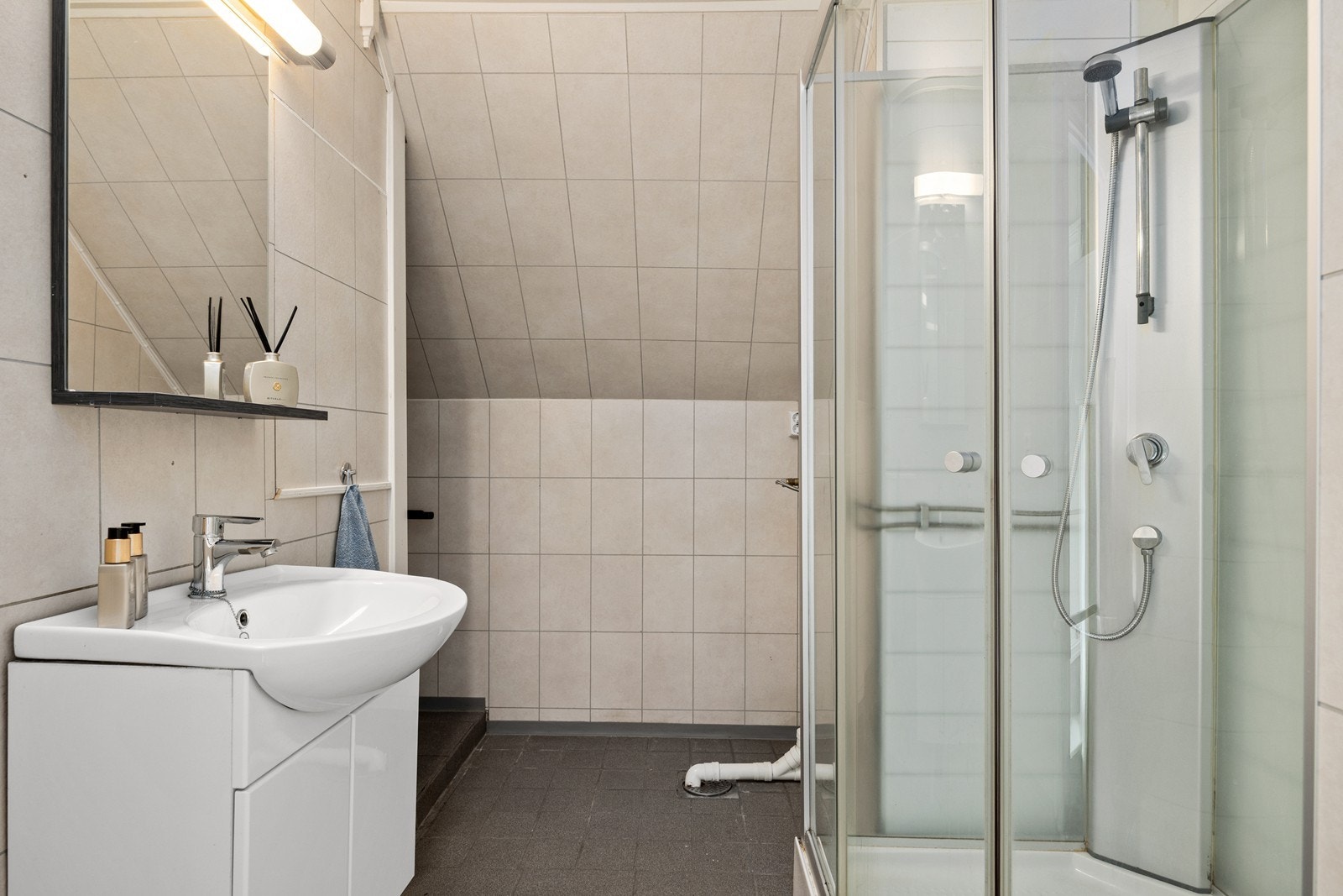 Bad med flislagt gulv og baderomsplater på vegger. Utstyrt med servantskap, dusjkabinett, wc på sokkel og opplegg for vaskemaskin. Galleribilde