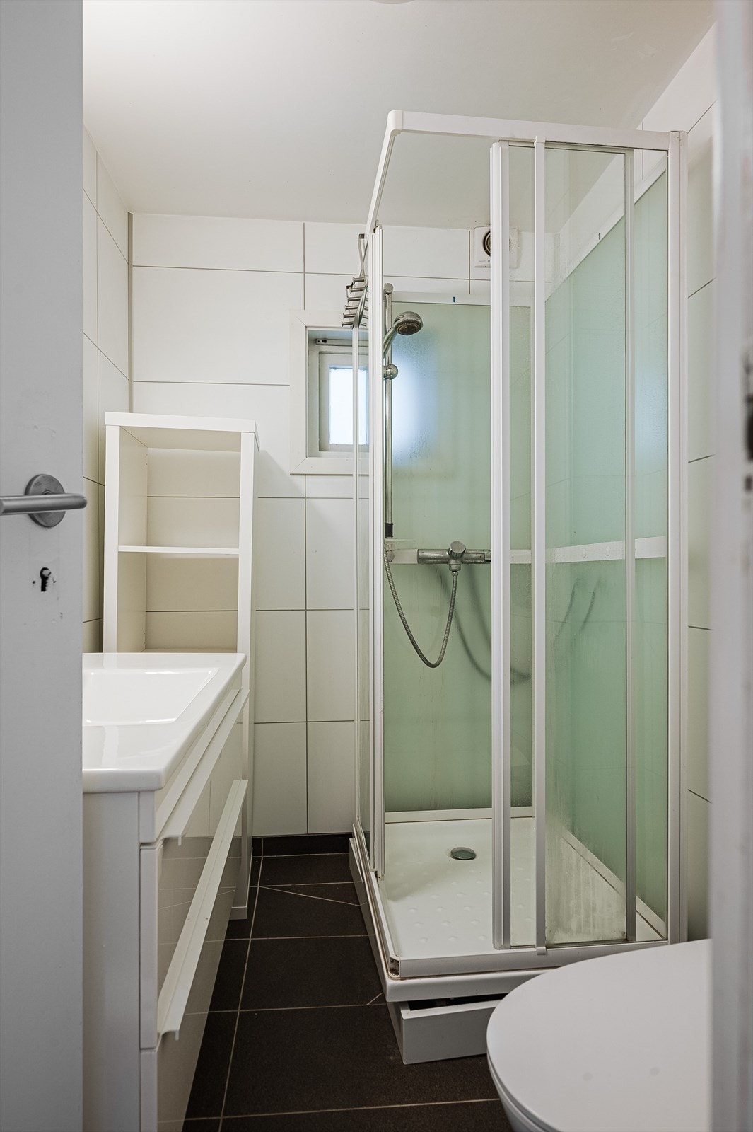 Badet har et klassisk preg med brune gulvfliser og hvite veggfliser. Varmkabler under gulvfliser gir et lunt baderom. Innredning med heldekkende servant med speil over, samt wc og dusjkabinett. Galleribilde