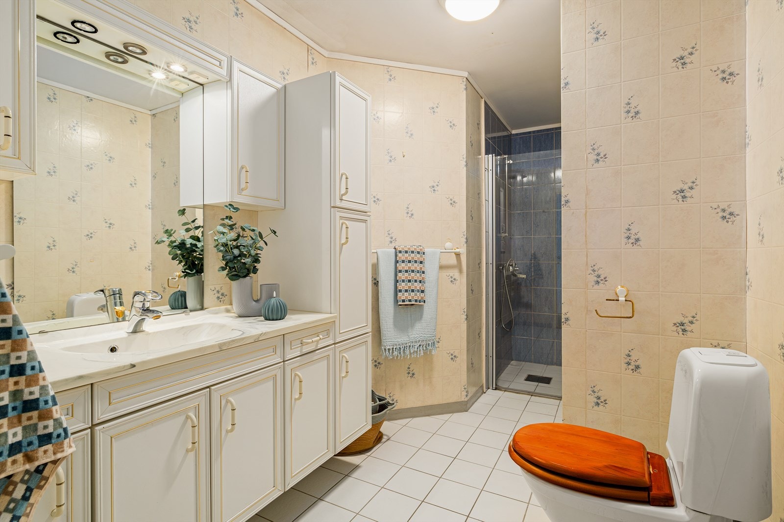 Romslig eldre bad med fliser på gulv og tapet på veggene. Servant i eldre, men innholdsrik innredning. Toalett. Galleribilde