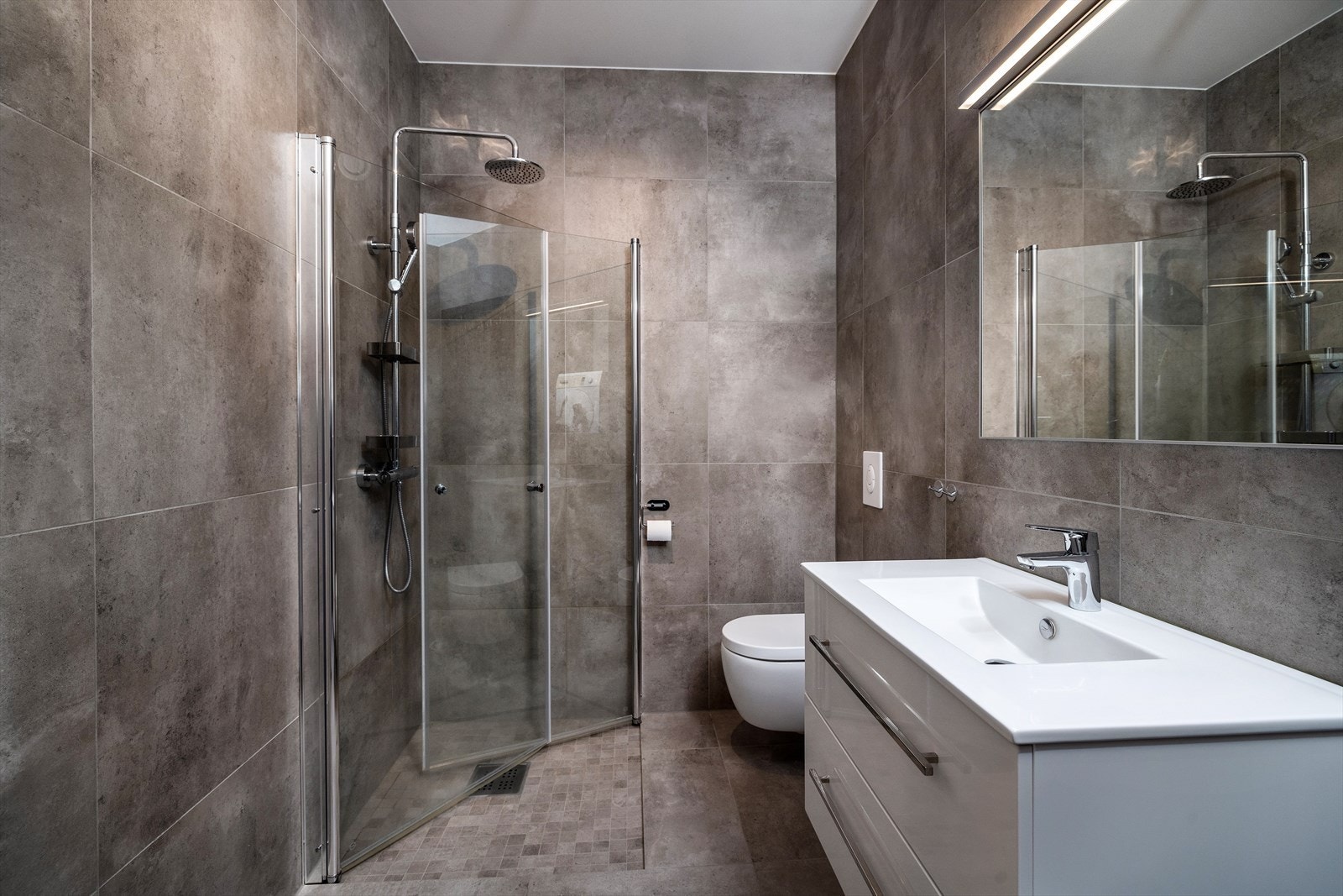 Tidsriktig innredet bad med elektrisk gulvvarme. Badet er utstyrt med dusjhjørne, vegghengt toalett, heldekkende nedfelt servant med rom for oppbevaring i skuffeseksjon og speil. Rommet har opplegg for vaskemaskin. Galleribilde