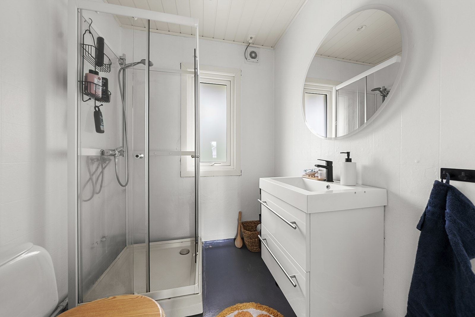 Baderom med belegg på gulv og malt våtromstapet på vegger. Rommet inneholder heldekkende servant over skuffeinnredning, overliggende speil med belysning, dusjkabinett og toalett Galleribilde