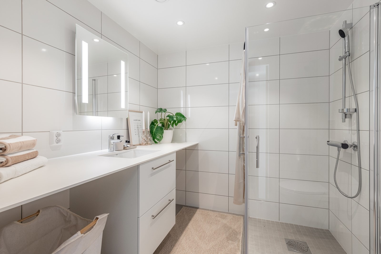 Romslig og flislagt baderom med varmekabler på gulv. Vaskemaskin og tørketrommel kan enkelt og plassbesparende plasseres under heldekkende benkeplate Galleribilde