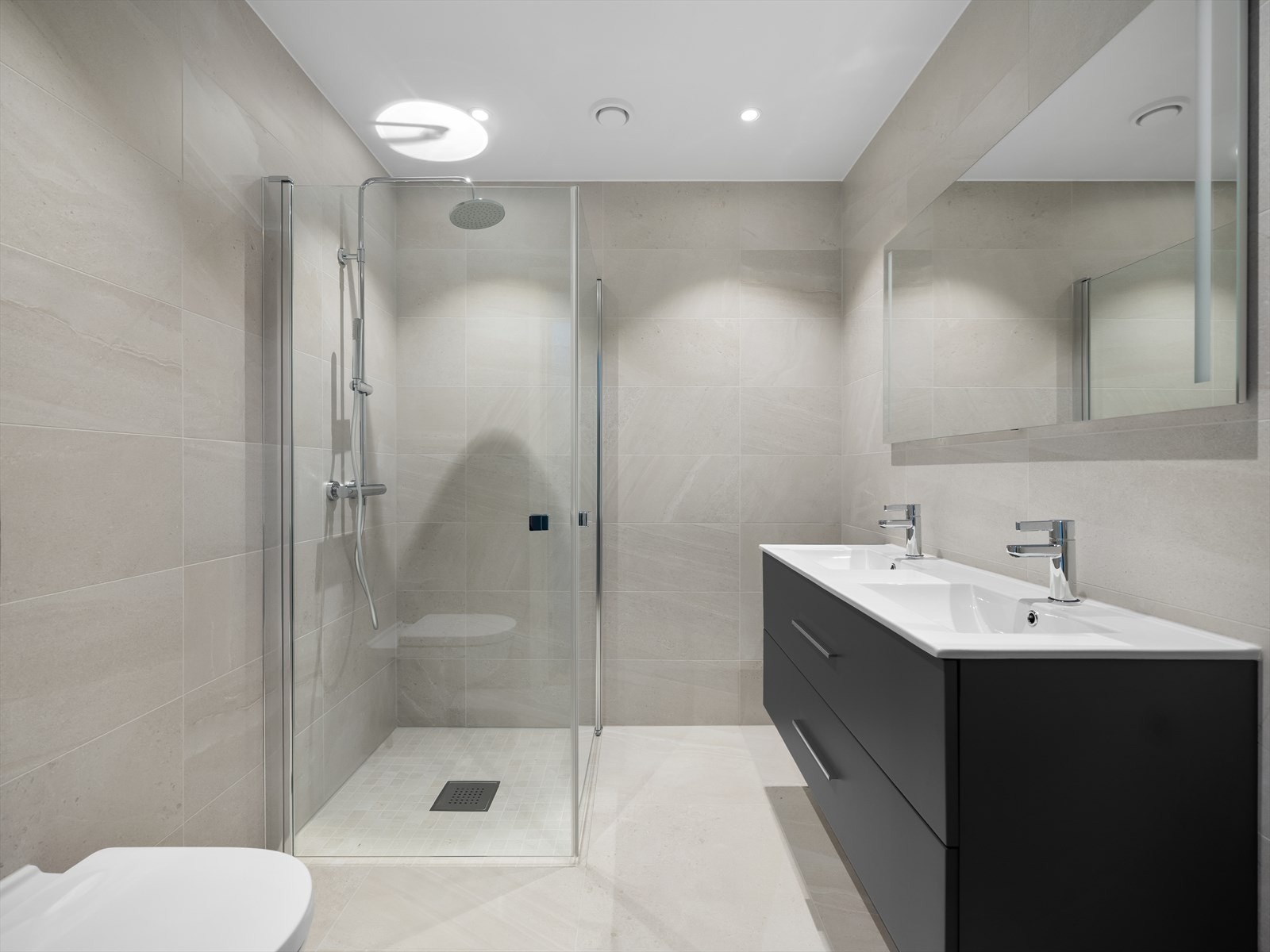 Lekre overflater med 60 x 60 Islanda, Nordic stone-fliser på gulvet og 30 x 60 mm fliser på veggene. Badet har moderne innredning fra Alterna med skuffer, servant og veggmontert speil med belysning. Galleribilde