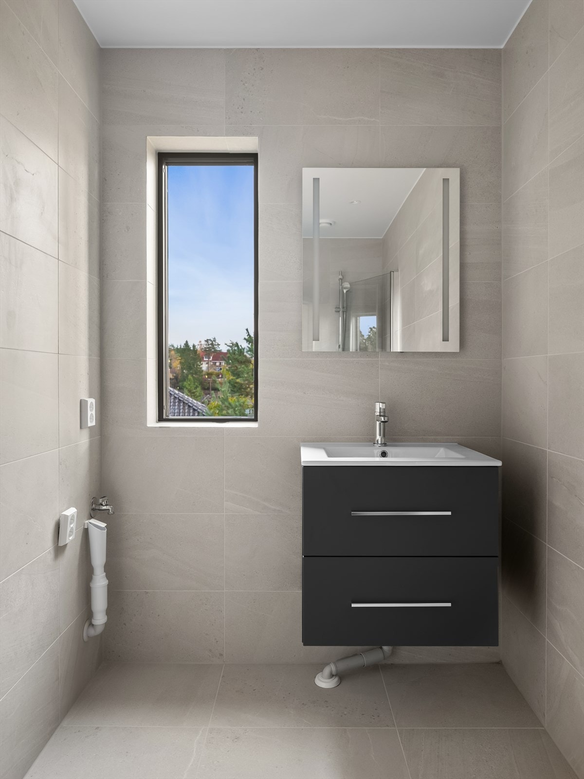 Badet har flislagte overflater og varmekabler. Rommet er utstyrt med toalett, dusjhjørne med innfellbare glassdører og baderomsinnredning fra Alterna med skuffer, servant og vegghengt speil. Vegghengt toalett og armatur fra Tapwell. Galleribilde