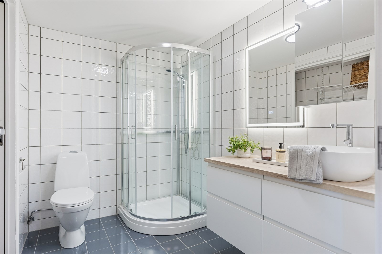 Lyst og moderne bad med flislagte vegger og gulv. Utstyrt med dusjkabinett, vegghengt speil med belysning og stilren innredning med servant. Galleribilde