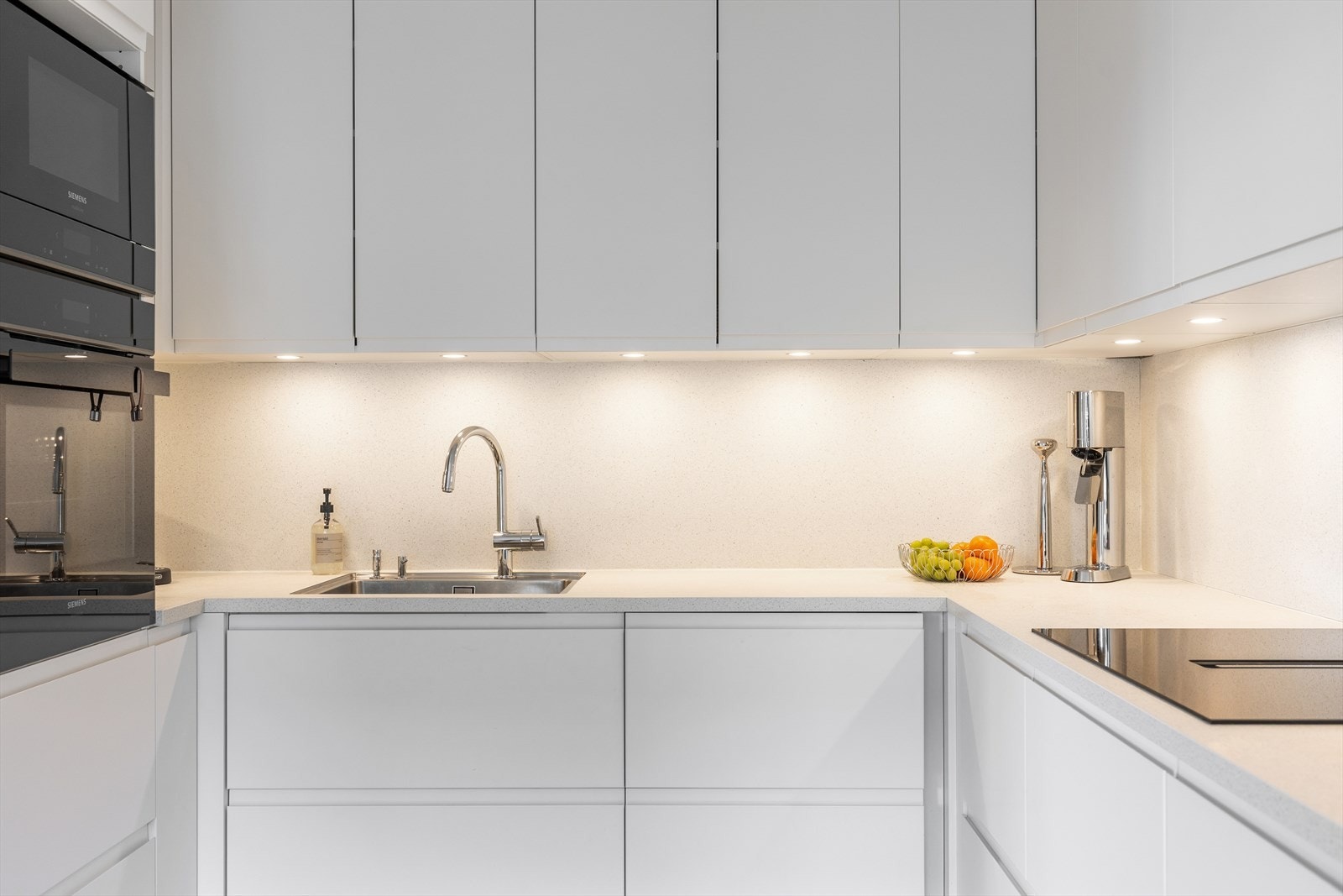 Kjøkkenet har glatte, stilrene fronter i lys utførelse som gir et rent og tidløst uttrykk. Benkeplaten er lys og harmonerer godt med resten av innredningen. Det er godt belyst med integrerte downlights under overskapene. Galleribilde