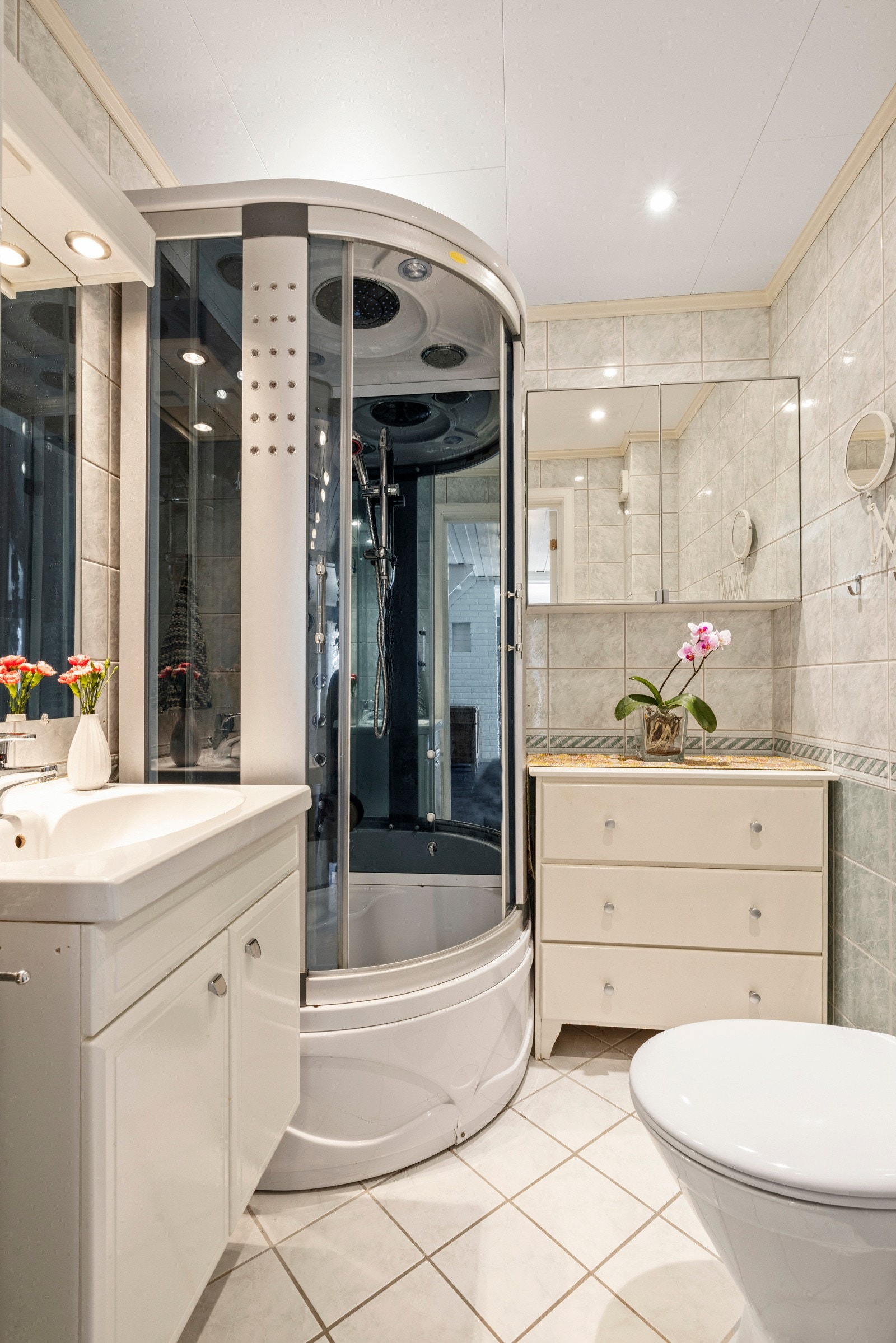 Badet i 1. etasje har flilagte overflater på gulv og vegger. Badet har toalett, dusjkabinett i herdet glass, speilskap, baderomsinnredning med høyglans fronter og heldekkende servant. Galleribilde