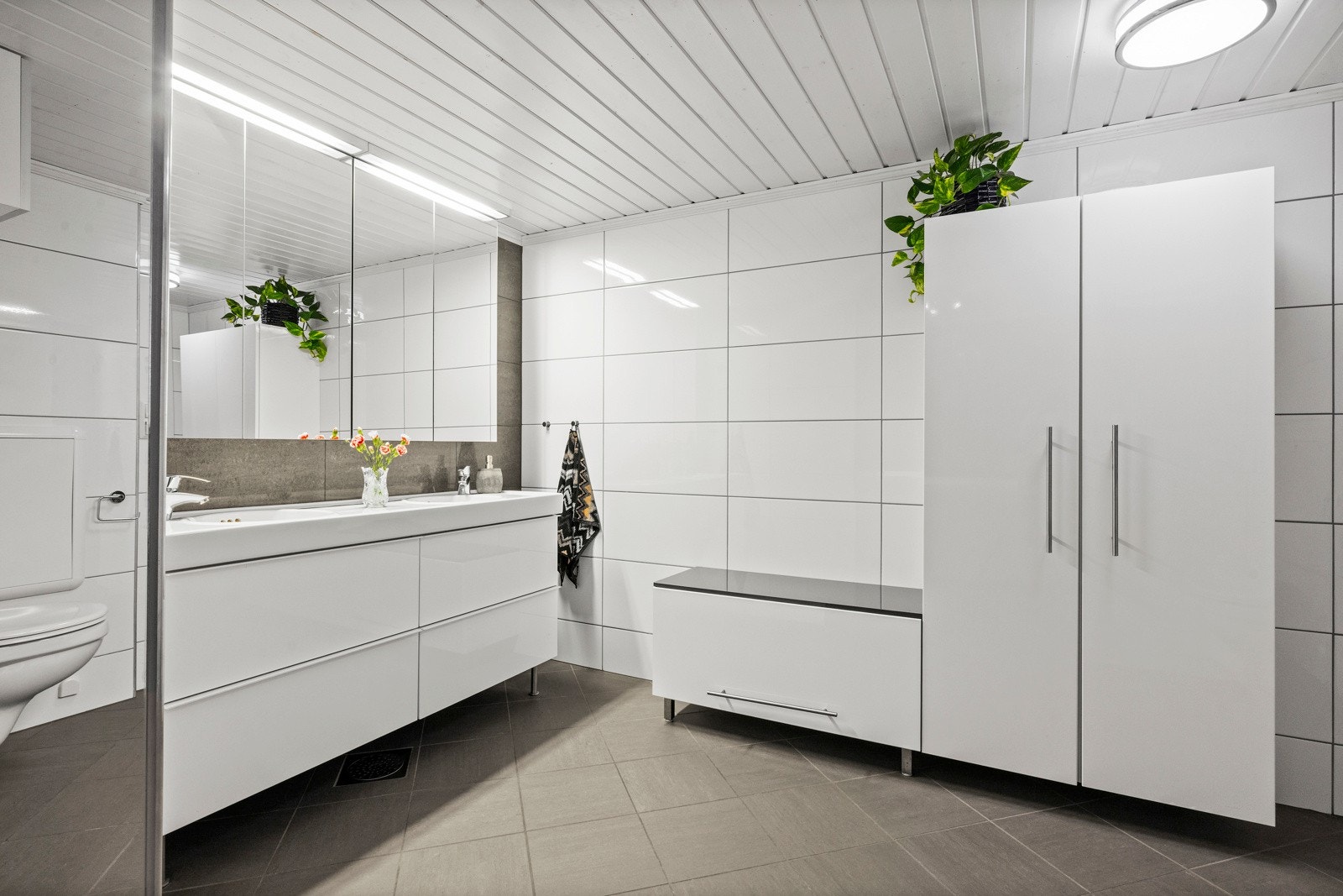 Badet er delt i to ulike soner. I den ene delen av rommet er det et veggmontert toalett, speilskap, skapinnredninger med høyglans fronter, baderomsinnredning med høyglans fronter og to heldekkende servanter. Galleribilde