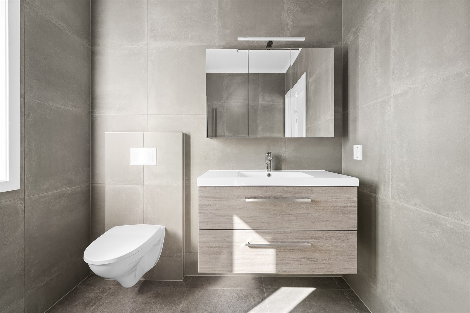 Badet er utstyrt med heldekkende servant over skuffeinnredning, speilskap med belysning, vegghengt toalett og dusjhjørne med innfellbare glassvegger Galleribilde