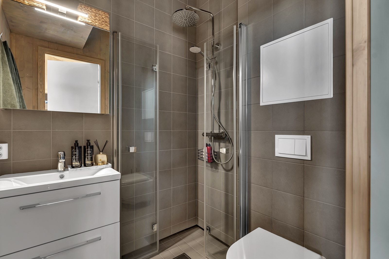 Badet er utstyrt med veggmonterte dusjdører, servant med skuffeseksjon, speil med belysning og veggmontert toalett Galleribilde