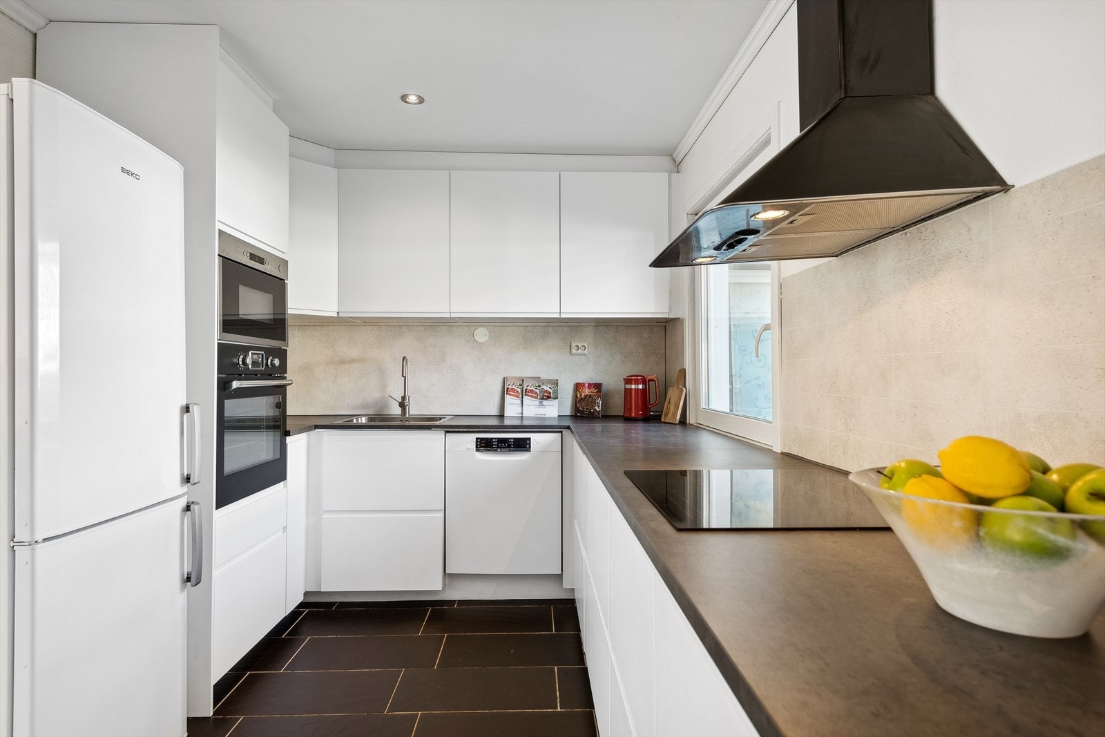 Kjøkkenet fra 2020 har glatte kjøkkenfronter, laminert benkeplate med nedfelt oppvaskkum, ventilator og dekorplate på vegg over benk. Galleribilde