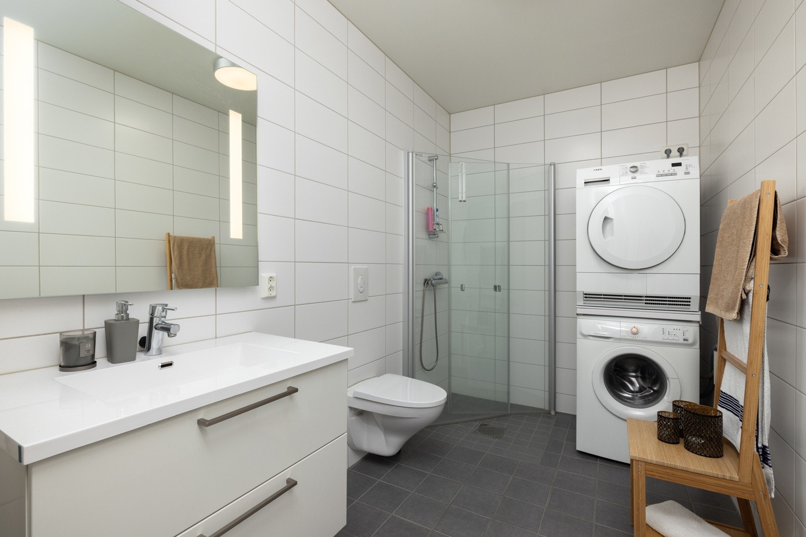 Fra entréen er det direkte adgang til ett av boligens to bad. Badet har flislagte gulv og vegger, samt elektriske varmekabler. Det er innredet med servant med ettgreps blandebatteri, veggmontert toalett, dusjhjørne med dusjvegger og opplegg for vaskemaski Galleribilde