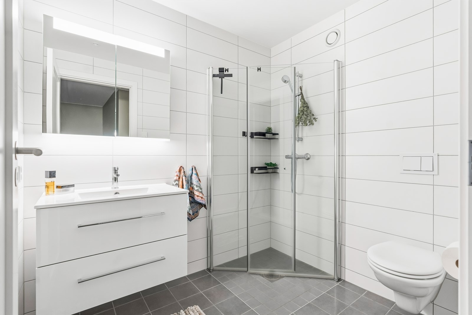 Delikat, flislagt baderom med dusjhjørne, vegghengt toalett, servantinnredning og speilskap. Galleribilde