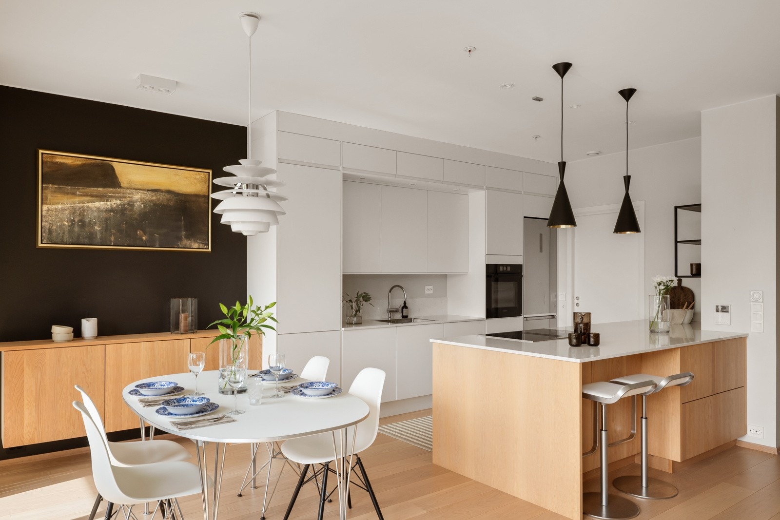 Innredningen er plassert langs én vegg med glatte, håndtaksløse fronter i hvitt, pluss en stor kjøkkenhalvøy som romdeler mot stuen. Galleribilde