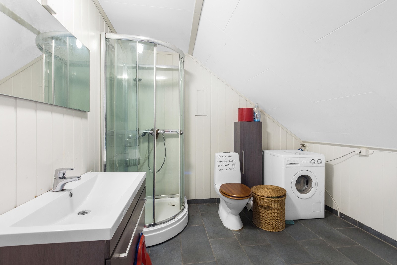 Badet har dusjkabinett, toalett, vaskemaskin og servant med speil. Veggene er panelt og gulvet har mørke fliser. Galleribilde
