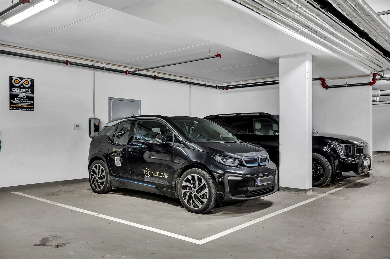 Det medfølger en romslig HC-parkeringsplass med elbillader, merket nr. 104. Boden ligger rett innenfor døren som vises i bakgrunnen, og er en egen, tilhørende bod. Galleribilde