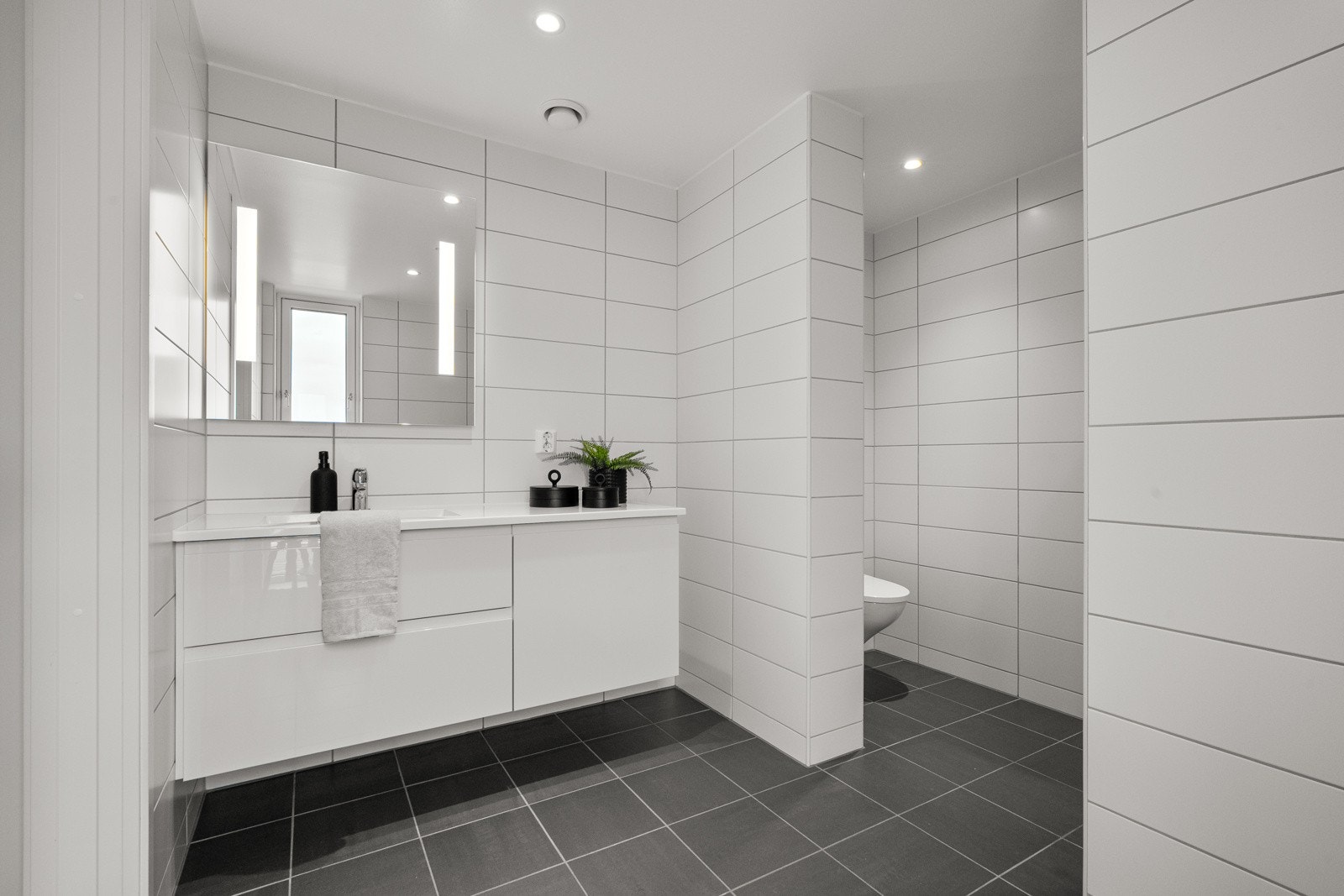 Baderommet har vegghengt servantskap med heldekkende servant, speil med lys og stikkontakt over servant, dusjnisje med glassdør og vegghengt toalett. Galleribilde