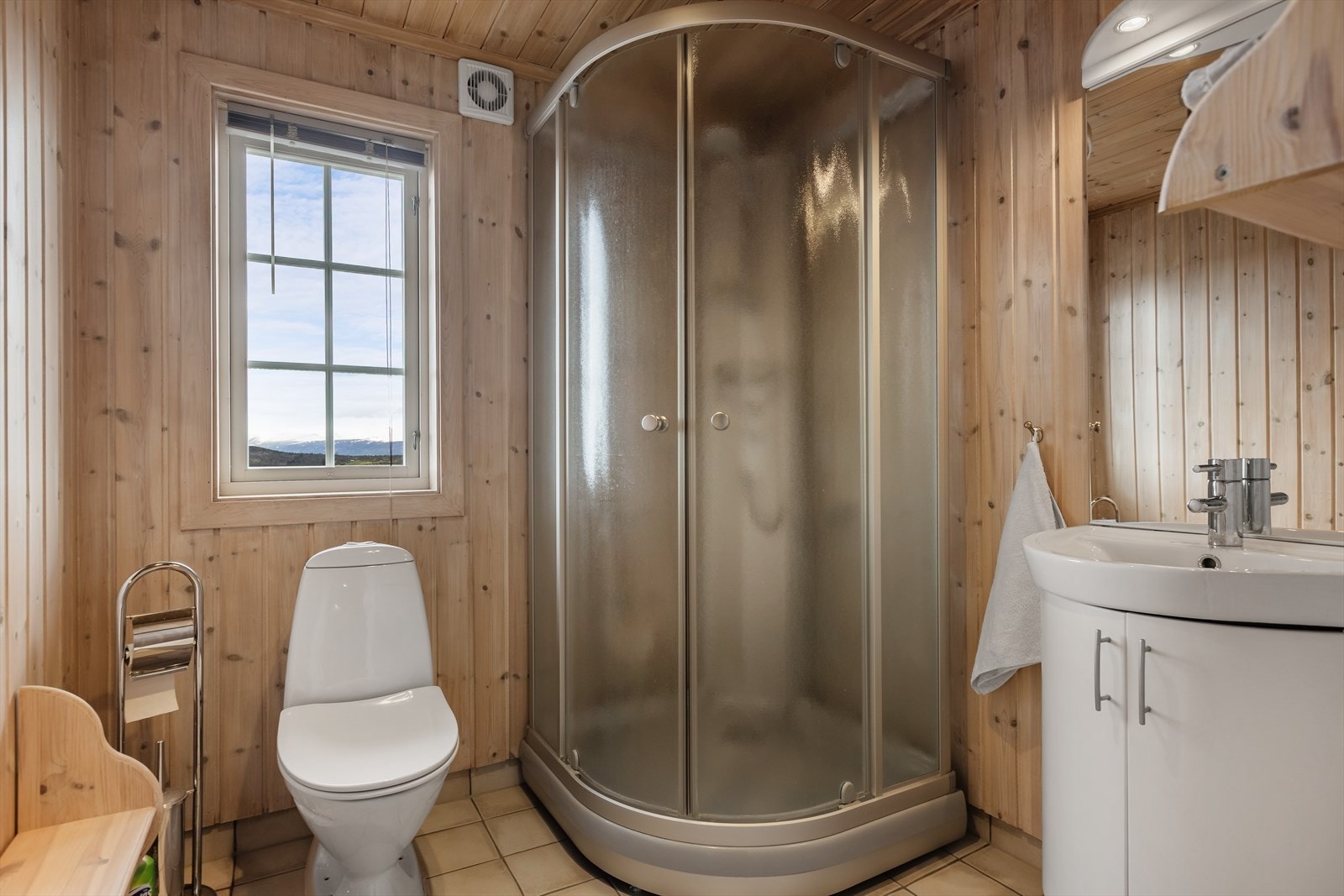 Bad har fliser med varmekabler på gulv, trepanel på vegg og himling. Klosett, servant og dusjkabinett. Galleribilde