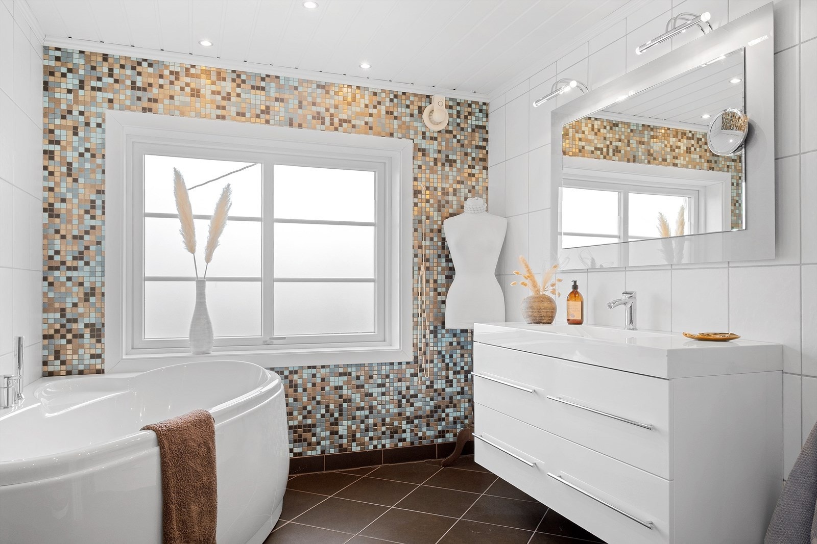 Badet har flislagte overflater med mosaikkfelt som gir et dekorativt uttrykk. Rommet har badekar, samt servantinnredning med god skapplass. Innfelte downlights i himling gir jevn belysning. Galleribilde