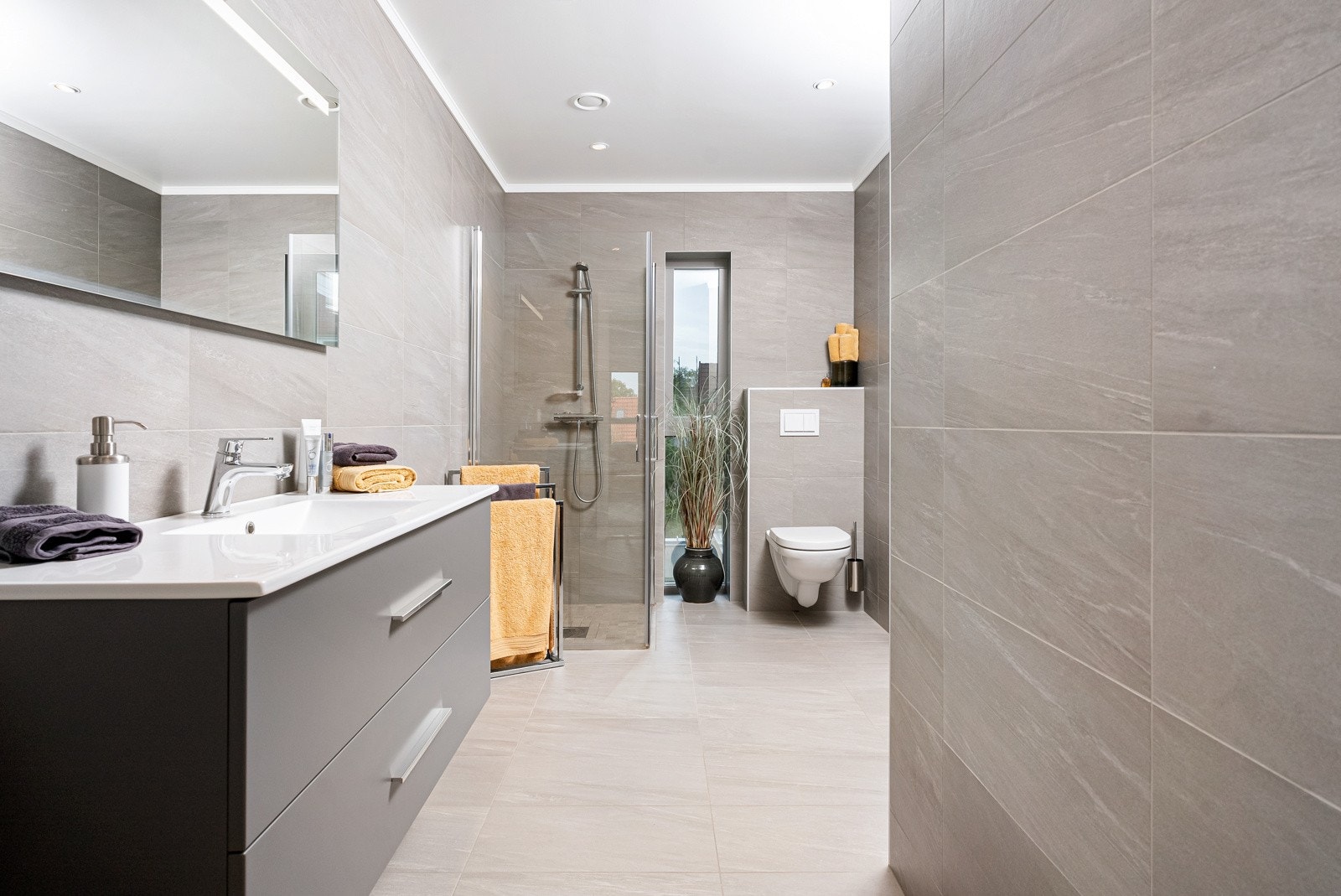 Begge badene har vegghengt toalett og et dusjhjørne med innfellbare dører, og baderomsinnredningen består av servant med grå skuffer og et speil med integrert belysning. Galleribilde