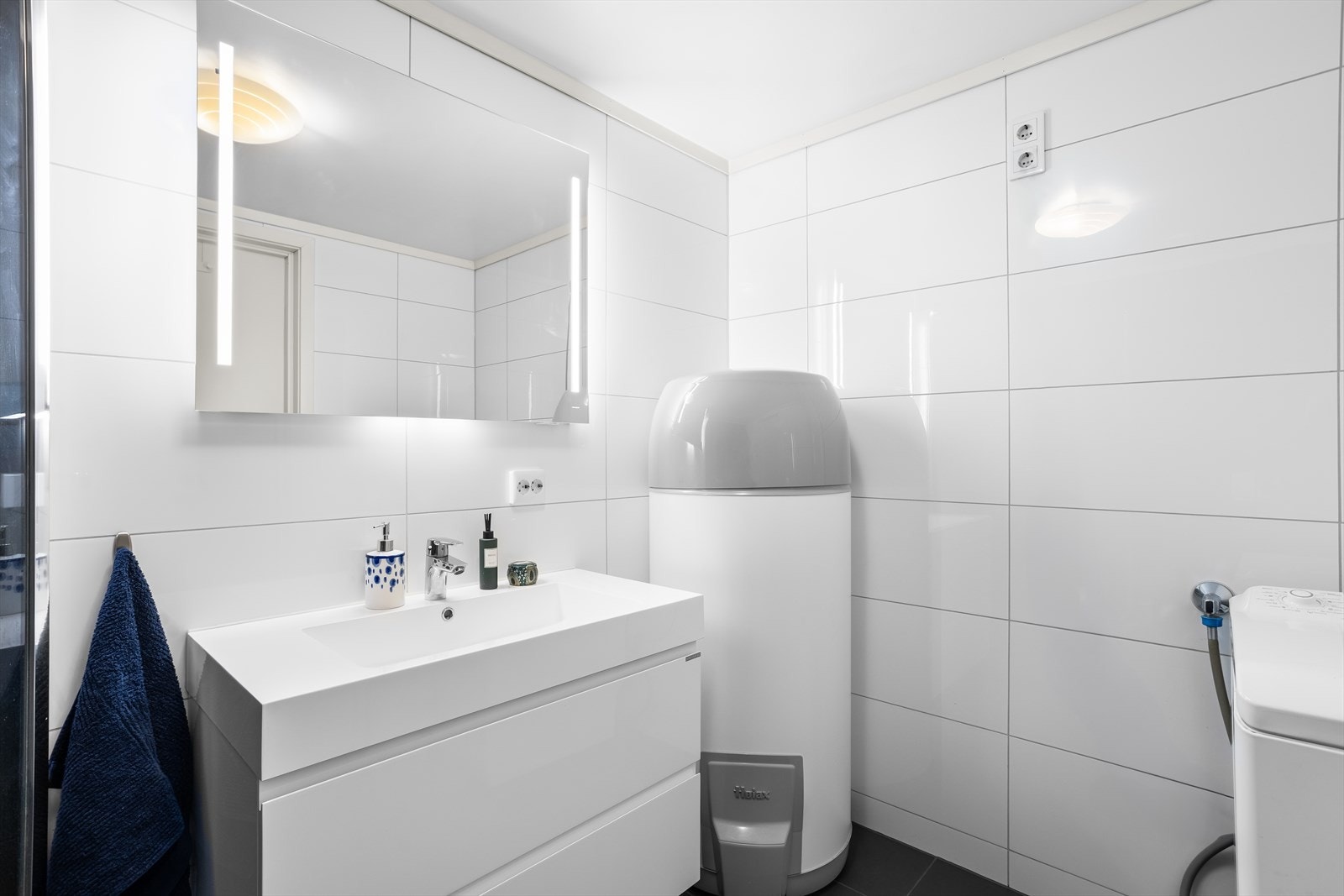 Badet er innredet med servant med underskap, speil med tilhørende belysning, vegghengt toalett, dusjdører og opplegg til vaskemaskin. Galleribilde